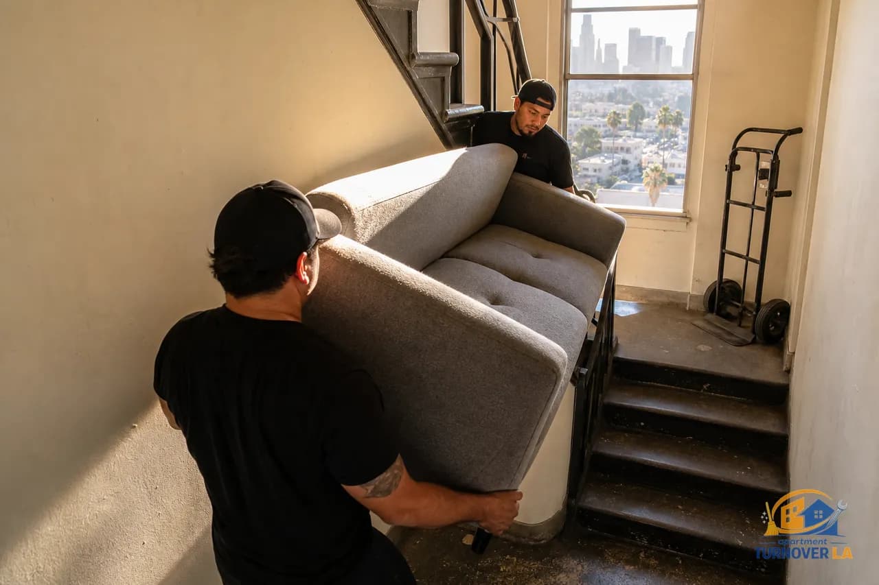 Haulers maneuver a sofa through a narrow LA apartment stairwell during furniture removal, with a faint Downtown skyline beyond the window